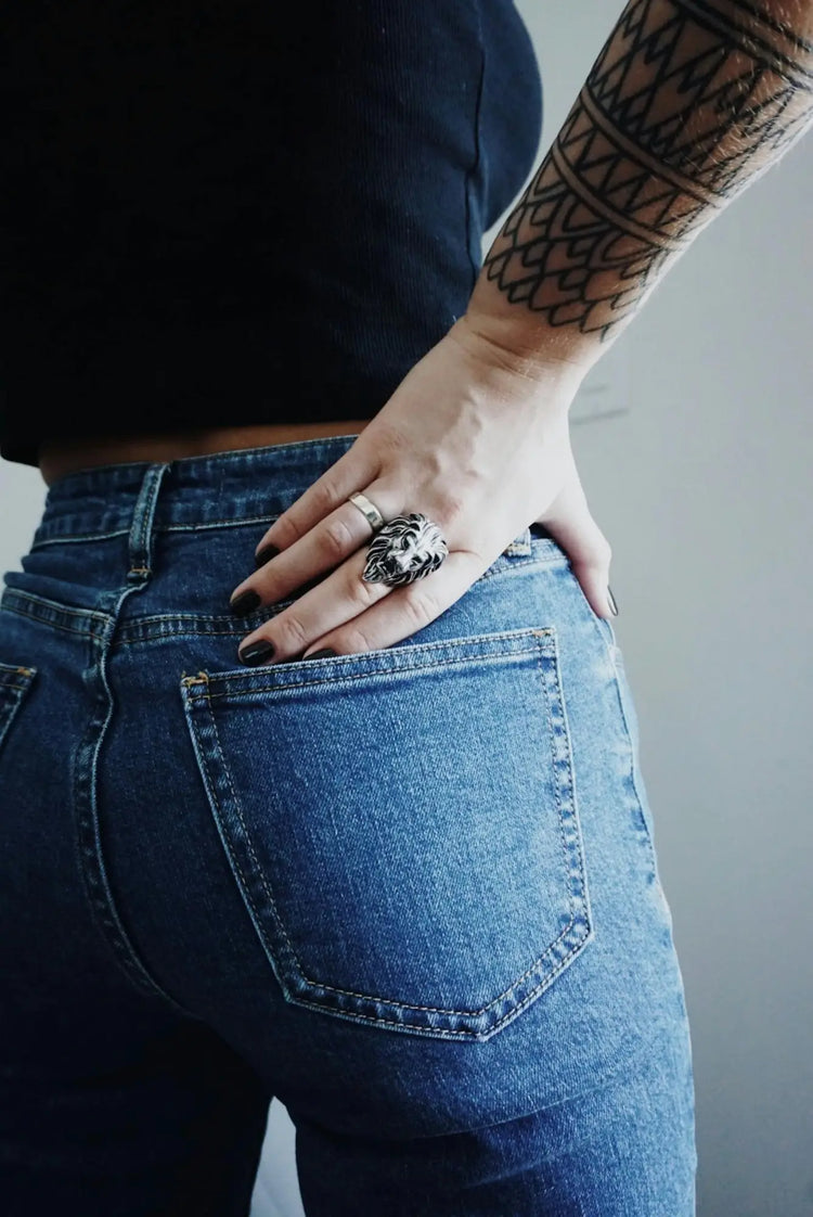 Hand with a decorative ring and tattoo resting on blue jeans pocket in Women’s Bottoms.